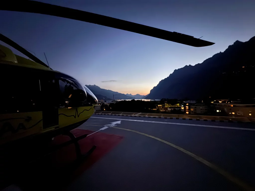 Rettungshubschrauber auf dem Landeplatz in der Abenddämmerung mit Bergpanorama im Hintergrund