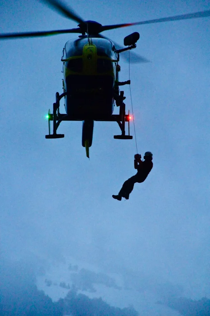 Rettungshubschrauber bei einer Windenrettung, eine Person hängt am Bergungsseil vor bewölktem Himmel